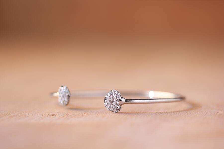 Una pulsera de plata con dos puntas redondas incrustadas de diamantes se exhibe sobre una superficie de madera clara, evocando la elegancia de los objetos de codicilo memorias testamentarias sobre un fondo marrón suave y difuminado.