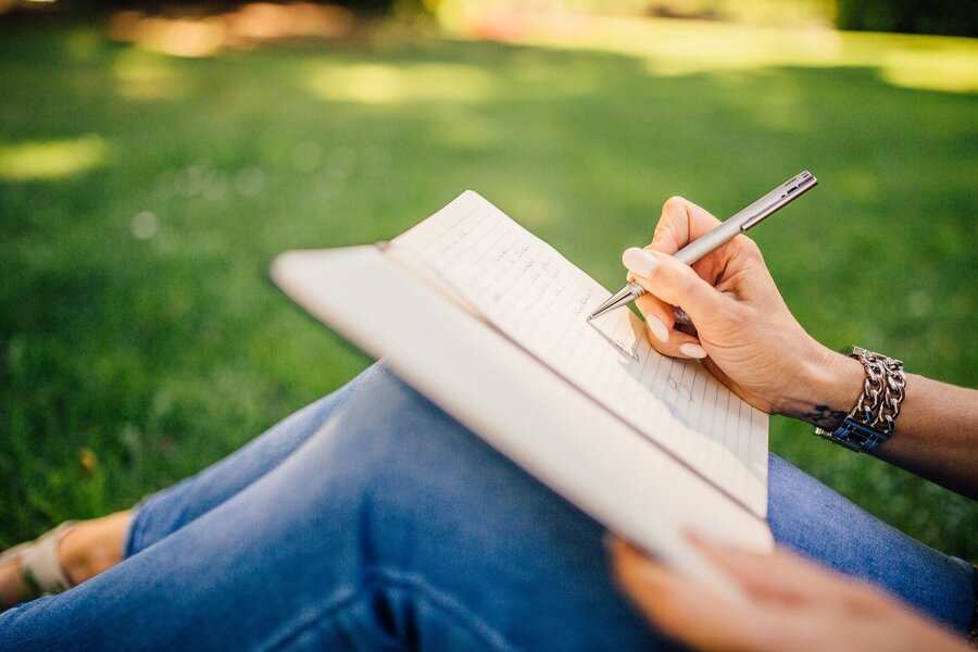 Una persona sentada en la hierba de un parque escribe codicilo memorias testamentarias en un cuaderno con un bolígrafo. Vestida con vaqueros azules y una pulsera, sólo se ven sus manos y su regazo sobre el fondo verde ligeramente difuminado.