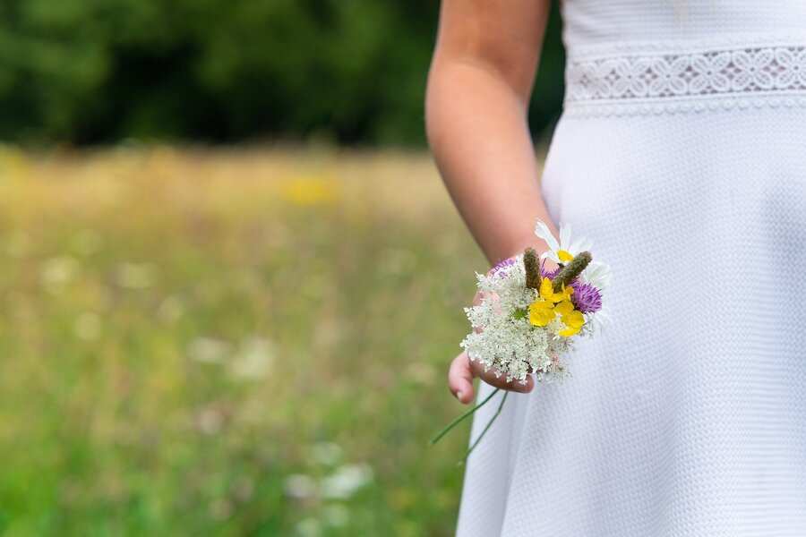 Una persona vestida de blanco está de pie en un campo, sosteniendo un pequeño ramo de flores silvestres: una imagen que evoca la inocencia de la primera comunión con padres divorciados. El fondo está difuminado con tonos verdes y amarillos, y no se ve el rostro. Una persona vestida de blanco está de pie en un campo, sosteniendo un pequeño ramo de flores silvestres: una imagen que evoca la inocencia de la primera comunión con padres divorciados. El fondo está difuminado con tonos verdes y amarillos, y no se ve el rostro.