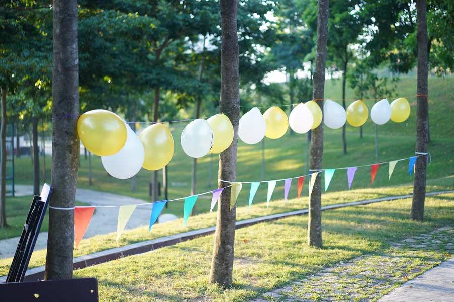 Una hilera de globos amarillos y blancos se ata entre los árboles de un parque para una primera comunión con padres divorciados. Debajo de los globos cuelgan banderines triangulares multicolores sobre la hierba verde y los árboles.