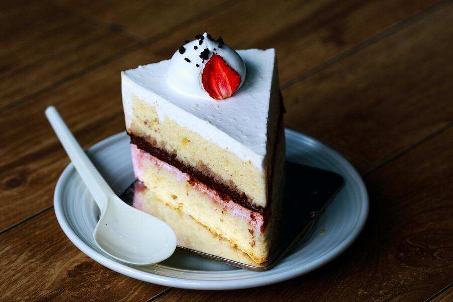 En un plato blanco con una cuchara de plástico se sirve un trozo de tarta con glaseado blanco, capas rosas y marrones, nata montada, un pequeño fruto rojo y chispas de chocolate, perfecta para celebrar una primera comunión con padres divorciados.