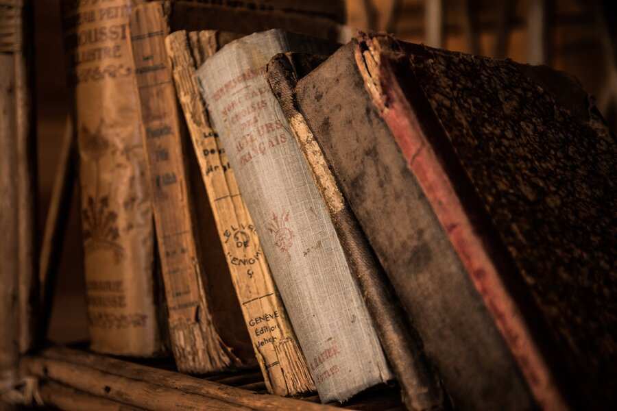 Varios libros viejos y desgastados con tapas y lomos descoloridos -quizá alguno oculte un testamento ológrafo olvidado- están alineados verticalmente en una estantería de madera, sus bordes deshilachados y superficies descoloridos revelan años de historia.