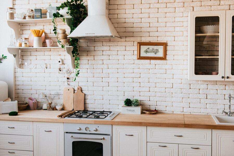Una cocina moderna con armarios blancos, una encimera de madera, cocina de gas, campana extractora, estantes abiertos con platos y plantas, tarros y un salpicadero de ladrillo, perfecta para el uso de vivienda familiar ¿hasta cuándo? Caja de pañuelos y tablas de cortar adornan el mostrador.