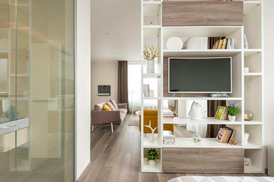 Moderno salón con suelo de madera clara, una estantería abierta blanca y de madera con libros, decoración y un televisor de pared. Uso vivienda familiar ¿hasta cuándo? A la izquierda hay una puerta de cristal, con un sofá beige y un gran ventanal al fondo. Moderno salón con suelo de madera clara, una estantería abierta blanca y de madera con libros, decoración y un televisor de pared. Uso vivienda familiar ¿hasta cuándo? A la izquierda hay una puerta de cristal, con un sofá beige y un gran ventanal al fondo.