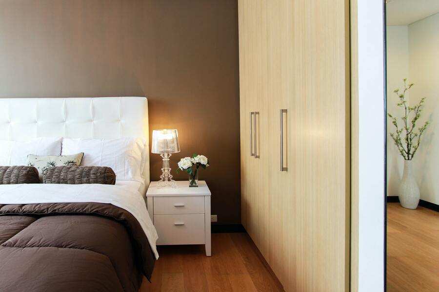 Un dormitorio moderno con cabecero blanco, ropa de cama marrón, mesilla de noche blanca con lámpara y flores, armario de madera clara y suelo de madera, ideal para uso de vivienda familiar ¿hasta cuándo? En una habitación contigua se ve un jarrón con ramas.
