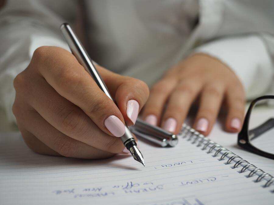 Una persona con las uñas pintadas de rosa claro escribe en un cuaderno de espiral sobre cómo cobrar pensión de viudedad tras un divorcio, utilizando una pluma estilográfica plateada. Las gafas y el capuchón del bolígrafo descansan cerca sobre el escritorio; se ve algo escrito.