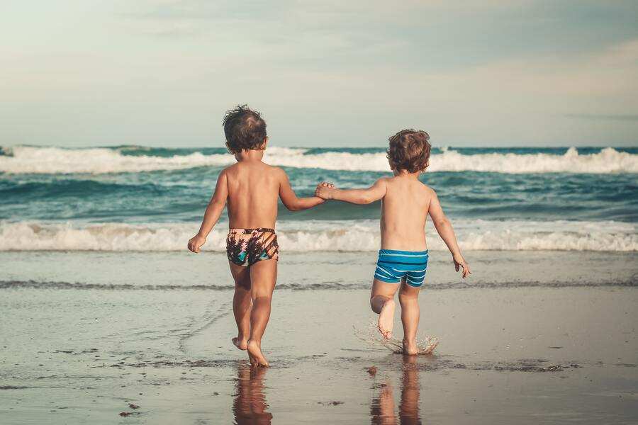 Dos niños pequeños en bañador, cogidos de la mano, caminan hacia el océano en una playa de arena con suaves olas de fondo bajo un cielo parcialmente nublado, disfrutando juntos de momentos despreocupados durante las vacaciones y la pensión de alimentos en verano.