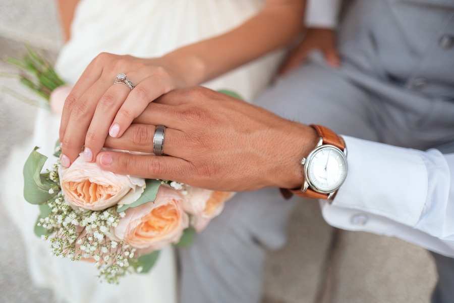 Primer plano de las manos de una pareja descansando sobre rosas de color rosa pálido; una mano muestra un anillo de diamantes, la otra una banda lisa y un reloj de pulsera. Ambos visten atuendo formal, lo que sugiere una boda ante notario o una ocasión especial. Primer plano de las manos de una pareja descansando sobre rosas de color rosa pálido; una mano muestra un anillo de diamantes, la otra una banda lisa y un reloj de pulsera. Ambos visten atuendo formal, lo que sugiere una boda ante notario o una ocasión especial.