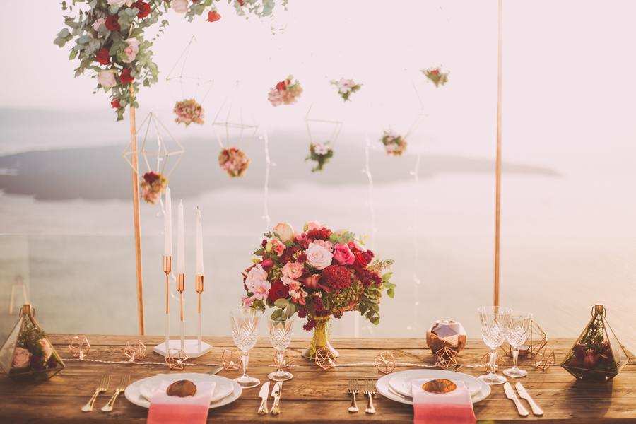 Una mesa de madera decorada para dos con platos, vasos, cubiertos, servilletas rosas, un arreglo floral central, velas y pequeños adornos geométricos, perfecta para una boda íntima ante notario con vistas al océano y flores colgantes al fondo.