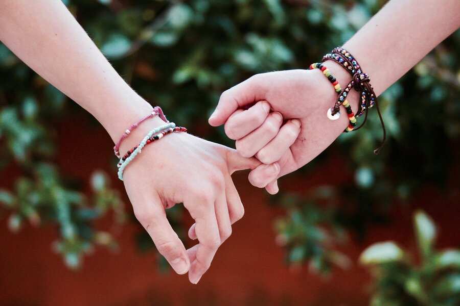 Dos manos con pulseras de cuentas de colores entrelazan sus dedos meñiques, simbolizando la unidad -muy parecida a los fuertes lazos que se ven en las herencias en parejas de hecho- sobre un fondo exterior difuminado con exuberante follaje verde.