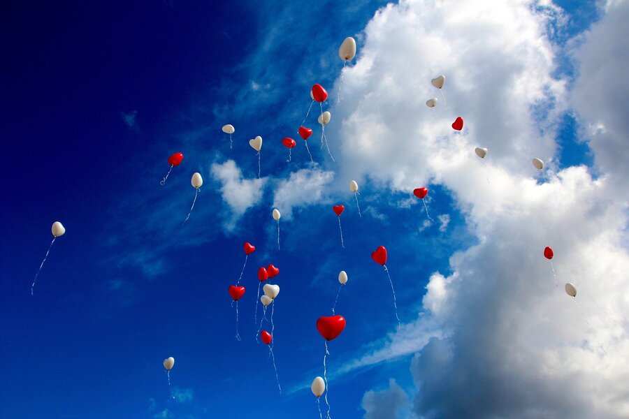 Un grupo de globos rojos en forma de corazón y blancos ovalados flotan hacia arriba sobre un cielo azul brillante con nubes dispersas, simbolizando el amor y la unidad, como las herencias en parejas de hecho.