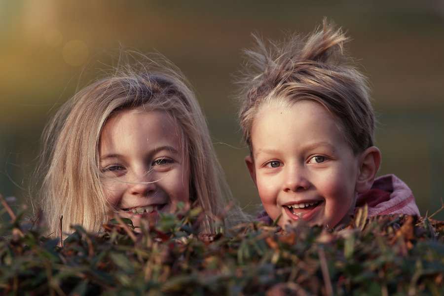 Dos niños sonrientes con el pelo claro, símbolos de la coparentalidad, se ven desde detrás de un seto con luz natural. La niña de la izquierda y el niño de la derecha llevan el pelo revuelto y miran directamente a la cámara.
