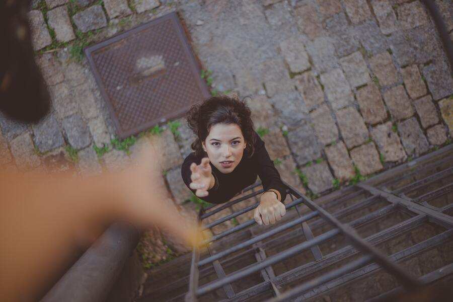 Una persona sube por una escalera metálica sujeta a una pared de ladrillo, tendiendo la mano hacia otra que la ayuda desde arriba.