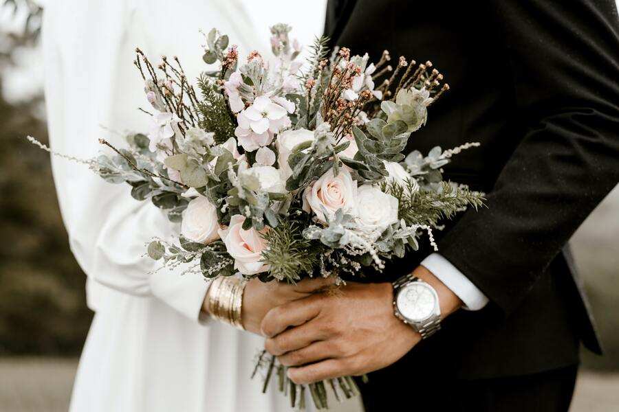 Una persona vestida de blanco y otra de traje negro, de pie y muy juntas, sostienen un gran ramo de flores rosas y blancas con vegetación, que representa la alegría tras cumplir los requisitos para contraer matrimonio. La persona del traje lleva un reloj de pulsera. Una persona vestida de blanco y otra de traje negro, de pie y muy juntas, sostienen un gran ramo de flores rosas y blancas con vegetación, que representa la alegría tras cumplir los requisitos para contraer matrimonio. La persona del traje lleva un reloj de pulsera.