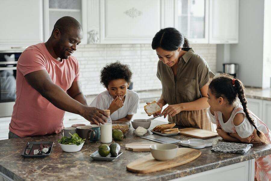 Un hombre, una mujer y dos niños preparan comida juntos en una cocina moderna, hablando de los requisitos para contraer matrimonio mientras están de pie alrededor de una isla de cocina con varios ingredientes y utensilios esparcidos.