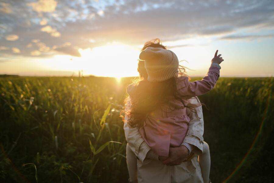 Un adulto lleva a un niño en brazos mientras, de pie en un campo cubierto de hierba al atardecer, reflexiona sobre los requisitos para contraer matrimonio. El niño, con sombrero, señala hacia el cielo mientras las nubes se deslizan sobre el sol bajo en el horizonte.