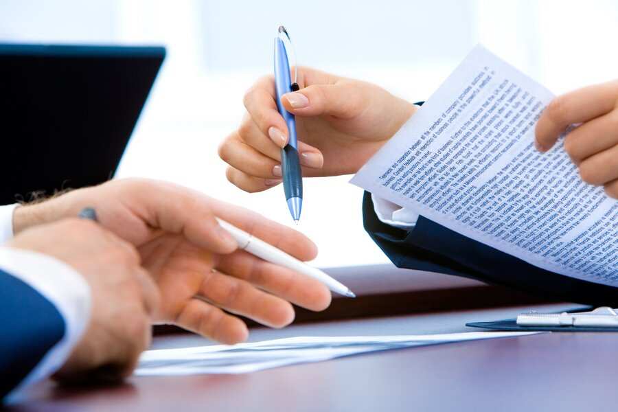 Dos personas en traje de negocios sosteniendo bolígrafos y un documento sobre despido disciplinario, sentadas en un escritorio con un ordenador portátil y papeles al fondo.