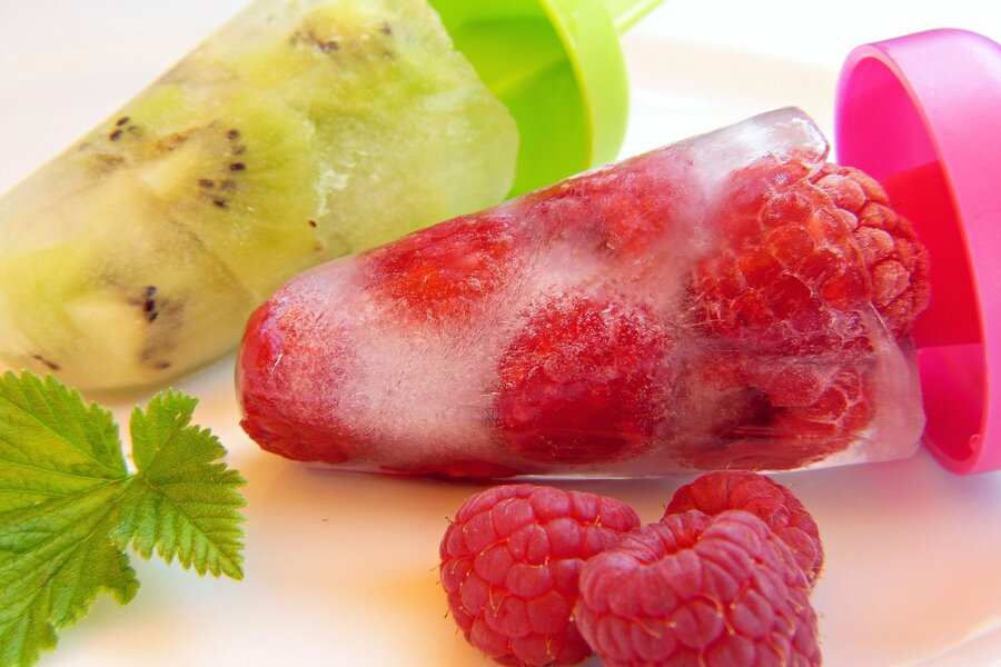 Dos polos de hielo caseros en un plato blanco; uno contiene frambuesas enteras en hielo transparente con un asa rosa, el otro tiene trozos de kiwi en hielo verde pálido con un asa verde. Cerca hay frambuesas frescas y una hoja verde, representan tanto el verano como la sensación que te queda cuando la empresa te deniega las vacaciones estivales.