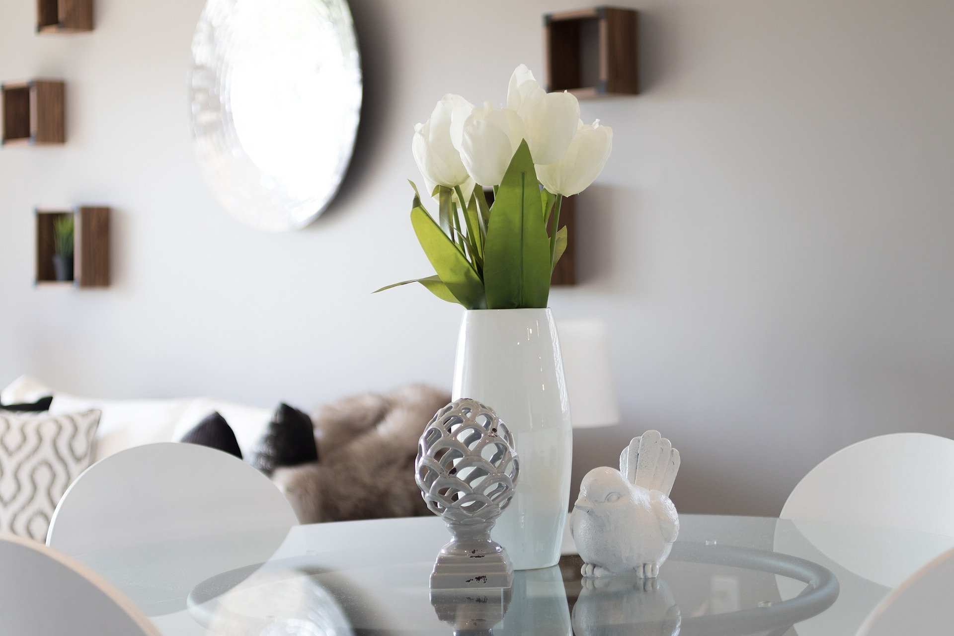 Un jarrón blanco con flores blancas descansa sobre una mesa redonda de cristal, acompañado de un pájaro de cerámica y un objeto decorativo. Al fondo, un sofá con cojines y unas estanterías de pared completan la escena, evocando tranquilidad, ideal para reflexionar sobre cuestiones de usufructo viudal.