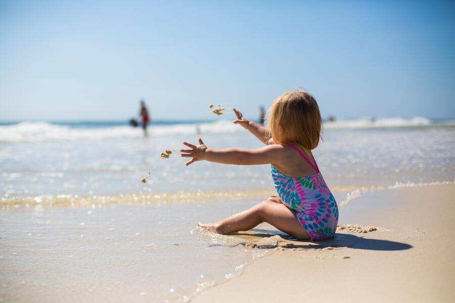 Un niño pequeño con un bañador de colores se sienta en la arena mojada de la playa, lanzando arena hacia el océano: un momento tranquilo de comunicación hijos durante las vacaciones con padres separados. El agua está en calma, con algunas personas al fondo cerca de la orilla.