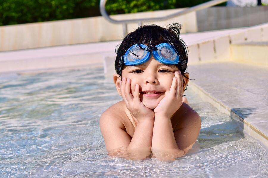 Un niño pequeño con gafas azules apoya la barbilla en las manos mientras se apoya en el borde de una piscina, sonriendo alegremente. El agua cristalina y el entorno tranquilo captan la alegría de las vacaciones, incluso para los hijos con padres separados. Escaleras y vegetación al fondo.