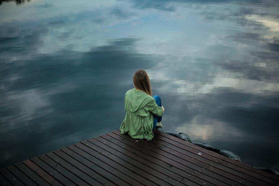 Una persona de pelo largo y chaqueta verde está sentada sola en un muelle de madera, mirando el agua tranquila y reflectante bajo un cielo nublado, sumida en sus pensamientos sobre los efectos del síndrome de alienación parental.