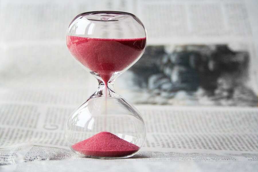 Un reloj de arena de cristal transparente con arena roja se coloca sobre un periódico, simbolizando cómo el tiempo mide tanto los retos como el progreso de las personas discapacitadas que se enfrentan a la incapacidad, a medida que la arena llena lentamente la cámara inferior.