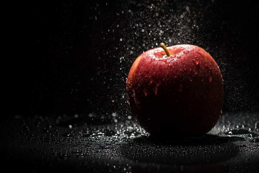 Una manzana roja con gotas de agua se asienta sobre una superficie oscura y húmeda sobre un fondo negro. La luz procedente de arriba resalta la manzana y la niebla que la rodea, simbolizando la frescura y la oportunidad para las personas discapacitadas que superan la incapacidad.