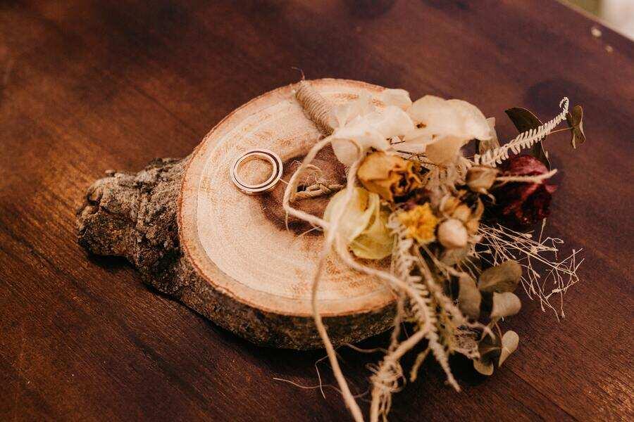 Dos anillos de oro descansan sobre una losa redonda de madera con bordes de corteza, junto a un ramo de flores secas, expuestas sobre una superficie de madera oscura, que evocan pensamientos sobre el viaje del amor e incluso momentos en los que la gente se plantea cómo divorciarse.