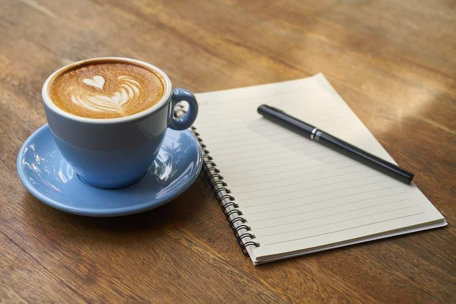 Sobre una mesa de madera, una taza de café con leche con espuma en forma de corazón reposa en un platillo azul junto a un cuaderno de espiral abierto y un bolígrafo negro, mientras alguien toma notas sobre cómo divorciarse. Sobre una mesa de madera, una taza de café con leche con espuma en forma de corazón reposa en un platillo azul junto a un cuaderno de espiral abierto y un bolígrafo negro, mientras alguien toma notas sobre cómo divorciarse.