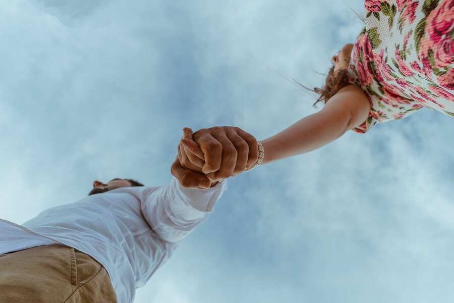 Vista en ángulo bajo de dos personas cogidas de la mano contra un cielo nublado, simbolizando la confianza, como pactos sucesorios. Una lleva camisa blanca y pantalones caqui; la otra, un vestido de flores. Sus rostros están parcialmente fuera de cuadro. Vista en ángulo bajo de dos personas cogidas de la mano contra un cielo nublado, simbolizando la confianza, como pactos sucesorios. Una lleva camisa blanca y pantalones caqui; la otra, un vestido de flores. Sus rostros están parcialmente fuera de cuadro.