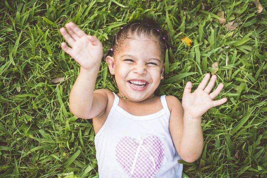 Un niño pequeño con una camiseta de tirantes blanca con un diseño de corazón está tumbado sobre hierba verde, sonriendo y levantando ambas manos en el aire. Con el pelo trenzado, el niño celebra haber terminado el pago campamento verano, irradiando pura felicidad.