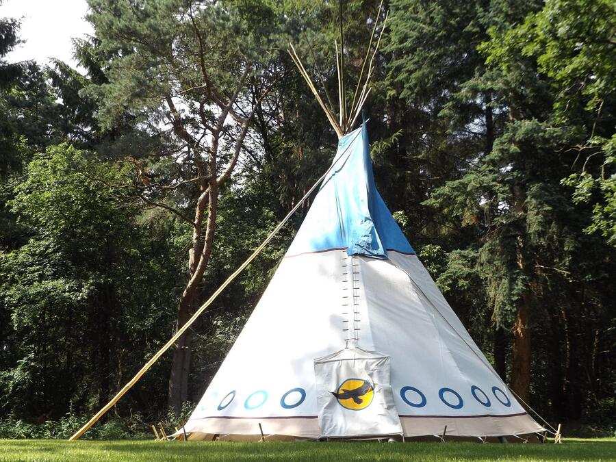 Un gran tipi blanco con detalles azules se alza sobre la hierba en un campamento de verano, rodeado de densos árboles verdes. El tipi tiene un pájaro pintado en la solapa delantera y varios palos altos que se extienden desde la parte superior.