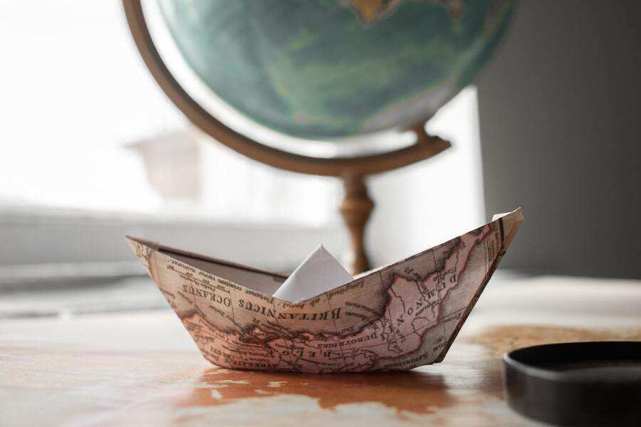 Un barco de papel hecho a partir de un mapa descansa sobre una mesa, evocando sueños de vacaciones con hijos en el extranjero. Un globo terráqueo se difumina en el fondo y parte de un objeto negro redondo se ve en primer plano.