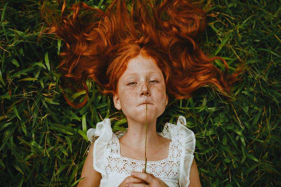 Una joven de larga melena pelirroja está tumbada sobre la hierba verde durante las vacaciones, con un vestido blanco de encaje. Sostiene un delgado tallo verticalmente delante de la cara, con el pelo extendido alrededor de la cabeza.