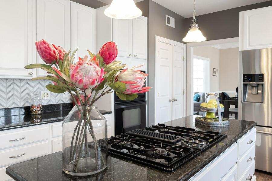 Una cocina moderna con armarios blancos, encimeras de granito negro, una placa de gas en la isla y electrodomésticos de acero inoxidable -perfecta para uso vivienda familiar- con un jarrón de flores rosas y lámparas colgantes colgando del techo. Una cocina moderna con armarios blancos, encimeras de granito negro, una placa de gas en la isla y electrodomésticos de acero inoxidable -perfecta para uso vivienda familiar- con un jarrón de flores rosas y lámparas colgantes colgando del techo.