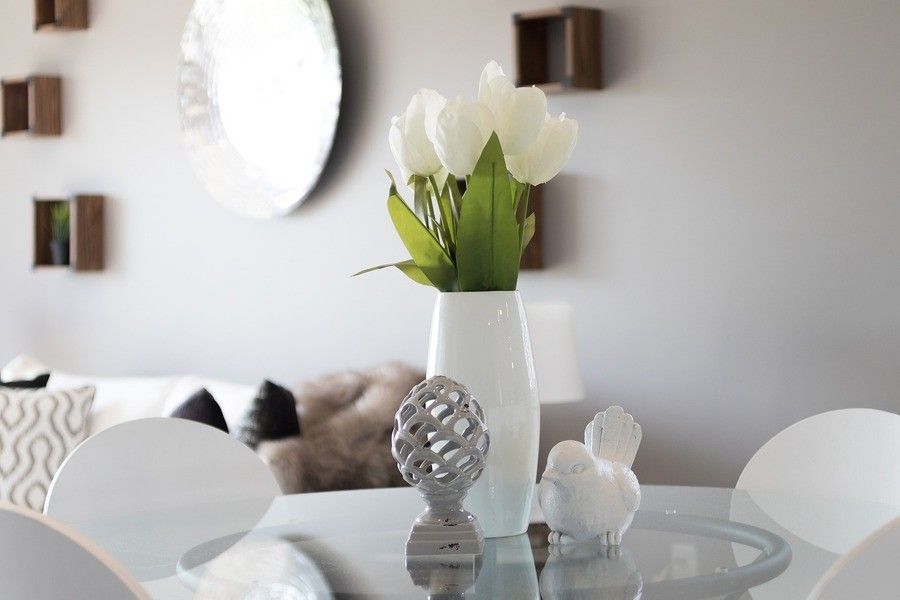 Un jarrón blanco con flores blancas descansa sobre una mesa de cristal, acompañado de un pájaro decorativo de cerámica y un adorno estampado. Al fondo, un sofá con cojines y sutiles toques de vivienda de estilo familiar.