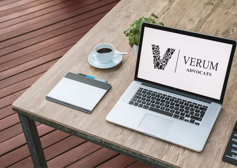 Un ordenador portátil con el logotipo de Verum Advocats sobre una mesa de madera, junto a una maceta, una tableta gráfica con bolígrafo y una taza de café.