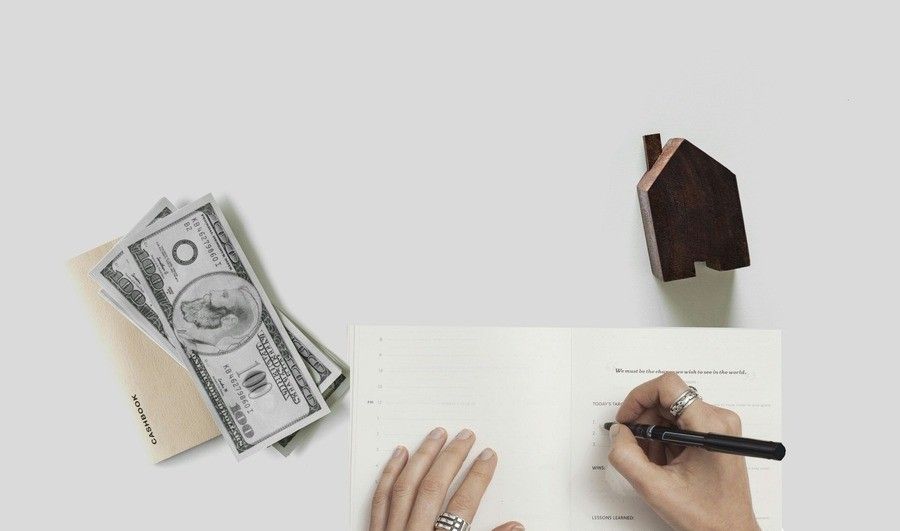 Una persona escribe en un cuaderno con un bolígrafo. Cerca, unos billetes de cien dólares estadounidenses y un pequeño objeto de madera con forma de casa descansan sobre una superficie blanca, sugiriendo asuntos relacionados con el divorcio contencioso y los arreglos financieros.
