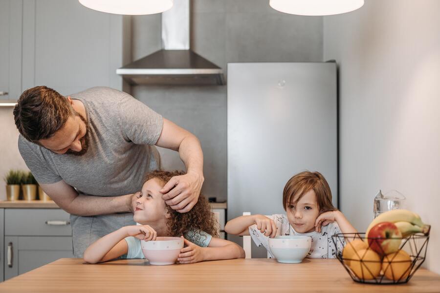 Un adulto, ejerciendo patria potestad y custodia, ayuda a un niño pequeño a arreglarse el pelo mientras otro niño se sienta a la mesa de la cocina. Ambos niños comen de cuencos en medio de una cocina con frigorífico, fogones y una cesta de fruta sobre la mesa.