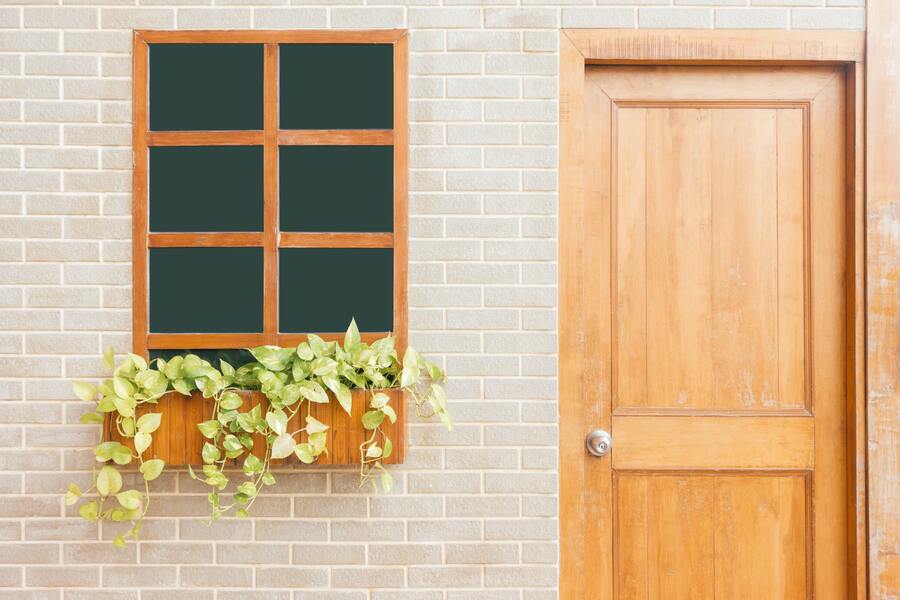 Una pared de ladrillo claro con una ventana de marco de madera dividida en seis cristales, una puerta de madera a la derecha y una jardinera de madera debajo de la ventana con plantas verdes de arrastre: una escena tranquila tocada por la realidad del desahucio por precario. Una pared de ladrillo claro con una ventana de marco de madera dividida en seis cristales, una puerta de madera a la derecha y una jardinera de madera debajo de la ventana con plantas verdes de arrastre: una escena tranquila tocada por la realidad del desahucio por precario.