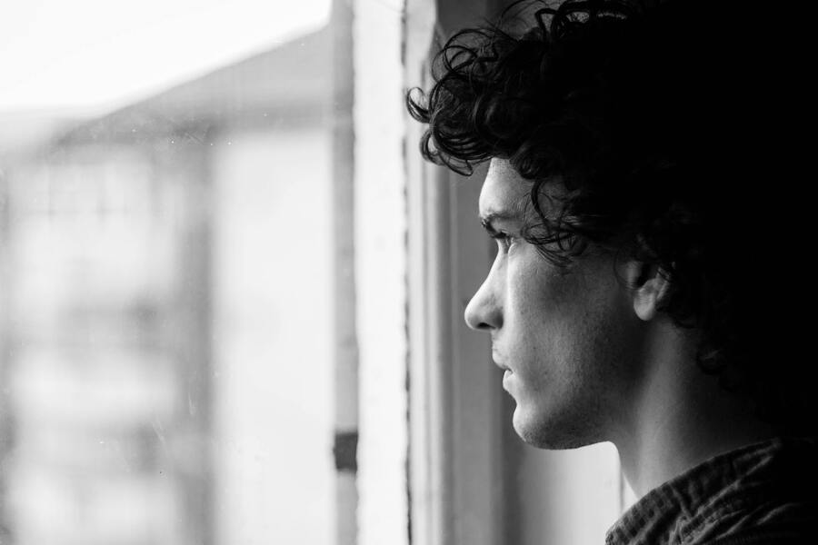Una persona con el pelo rizado mira por una ventana, su perfil enmarcado en blanco y negro. La imagen evoca una sensación de abandono hogareño familiar, ya que el fondo borroso insinúa recuerdos dejados atrás.