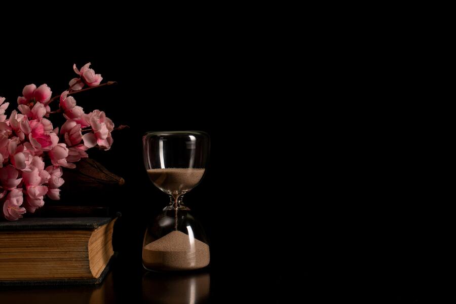 Un reloj de arena junto a un viejo libro con la inscripción RENUNCIA HERENCIA y un ramillete de flores rosas, todo ello sobre un fondo negro liso. Un reloj de arena junto a un viejo libro con la inscripción RENUNCIA HERENCIA y un ramillete de flores rosas, todo ello sobre un fondo negro liso.