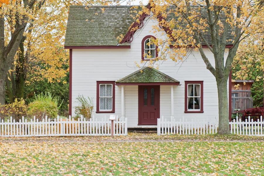 Una casa blanca de dos plantas con adornos rojos y una puerta de entrada roja, rodeada de una valla blanca y árboles otoñales. Perfecta para familias en situaciones como la del divorcio con hipoteca conjunta. Una casa blanca de dos plantas con adornos rojos y una puerta de entrada roja, rodeada de una valla blanca y árboles otoñales. Perfecta para familias en situaciones como la del divorcio con hipoteca conjunta.