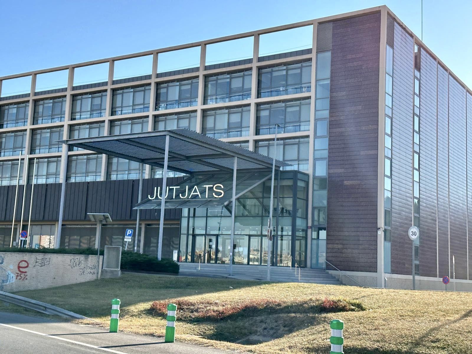 Un edificio moderno con grandes ventanales y un letrero que reza JUTJATS sobre la entrada, esta estructura -parte de los juzgados de Terrassa- cuenta con paneles de cristal, un toldo metálico y zonas de césped con señales de tráfico en primer plano. Un edificio moderno con grandes ventanales y un letrero que reza JUTJATS sobre la entrada, esta estructura -parte de los juzgados de Terrassa- cuenta con paneles de cristal, un toldo metálico y zonas de césped con señales de tráfico en primer plano.