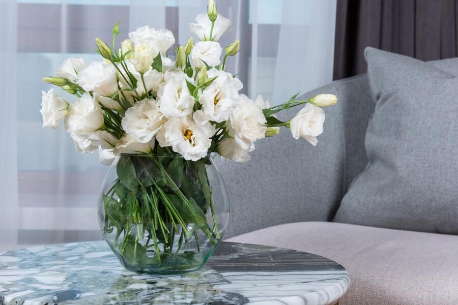 Un jarrón de cristal transparente lleno de flores blancas descansa sobre una mesa redonda de mármol junto a un sofá gris claro: un espacio elegante y tranquilo, perfecto para hablar de asuntos importantes como el divorcio con hipoteca conjunta. Unas cortinas transparentes y un cortinaje gris oscuro completan la escena.