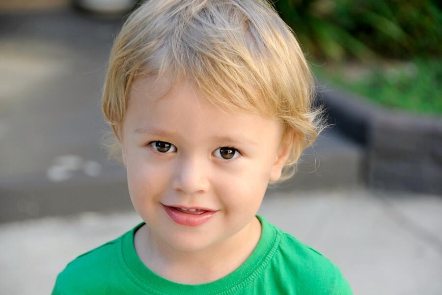 Convenio regulador de custodia compartida Un niño de pelo rubio claro y ojos marrones sonríe con una camisa verde brillante. Al aire libre, con el pavimento difuminado y la vegetación de fondo, la escena es tan armoniosa como un convenio regulador bien elaborado.