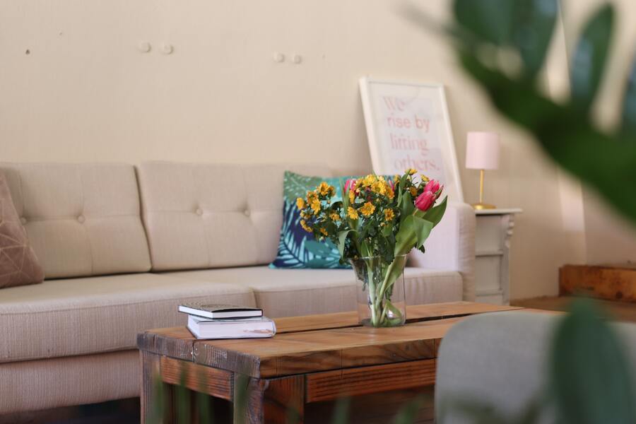 Un sofá beige con cojines se encuentra detrás de una mesa de centro de madera que sostiene un jarrón con flores de colores y dos libros sobre divorcio notarial. Al fondo, una cita enmarcada, una lámpara y una mesita. En primer plano se ven parcialmente hojas verdes.
