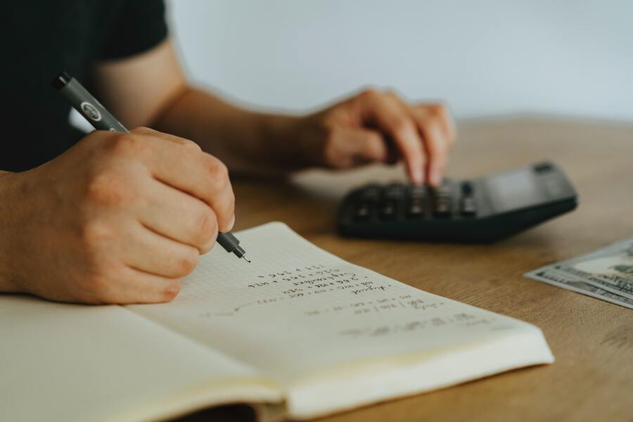 Una persona escribe cálculos en un cuaderno con una mano mientras utiliza una calculadora con la otra, posiblemente para declarar cobro herencia. Sobre la mesa de madera se ven algunos billetes de dólar estadounidense. Una persona escribe cálculos en un cuaderno con una mano mientras utiliza una calculadora con la otra, posiblemente para declarar cobro herencia. Sobre la mesa de madera se ven algunos billetes de dólar estadounidense.