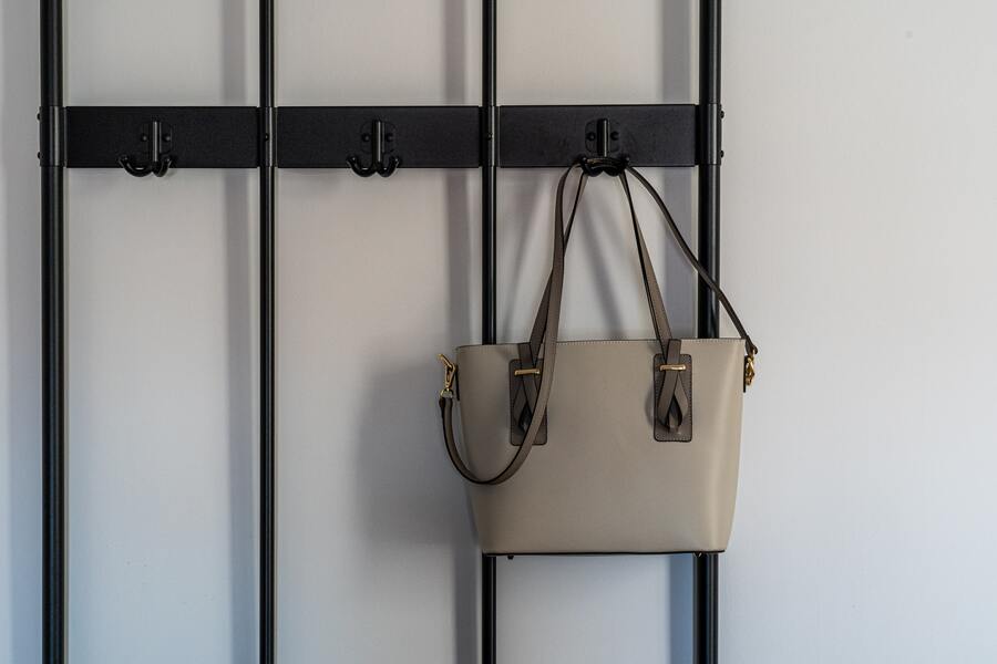 Un bolso beige con asas marrones, perfecto para las reuniones para declarar cobro herencia, cuelga de un perchero negro montado contra una pared blanca lisa.